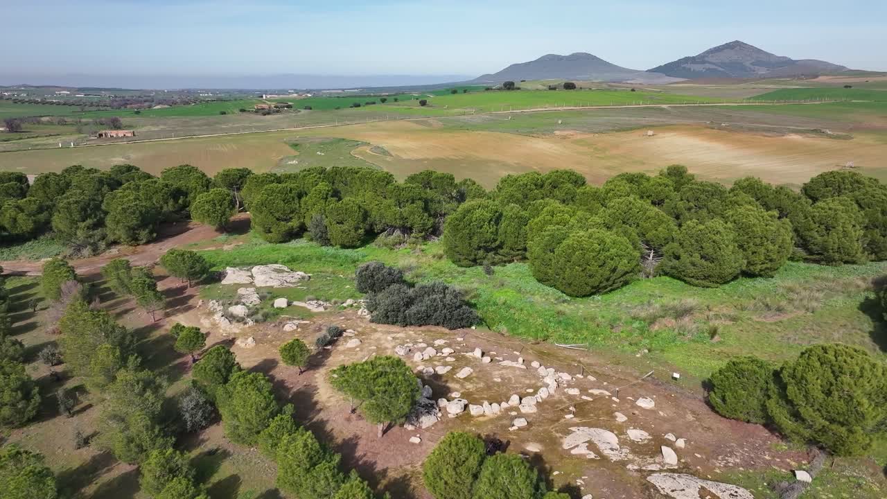 vuelo sobre un área de pinos y visualizamos un cromlech de piedras en un entorno de tierras de cultivo con un fondo de dos montañas con un cielo azul en una mañana de invierno en toledo, españa.