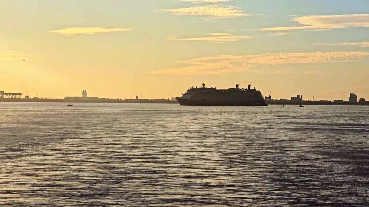 A wide scenic shot of a large cruise ship sailing across calm waters at sunset. The vessel and distant industrial coastal skyline form dark silhouettes against a vibrant golden yellow sky.