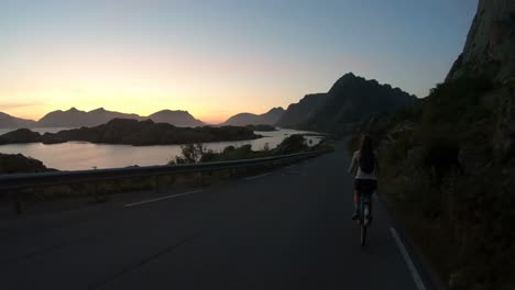 a young woman is biking down a hill on an old bicycle on the road near henningsvaer in lofoten during a beautiful sunset