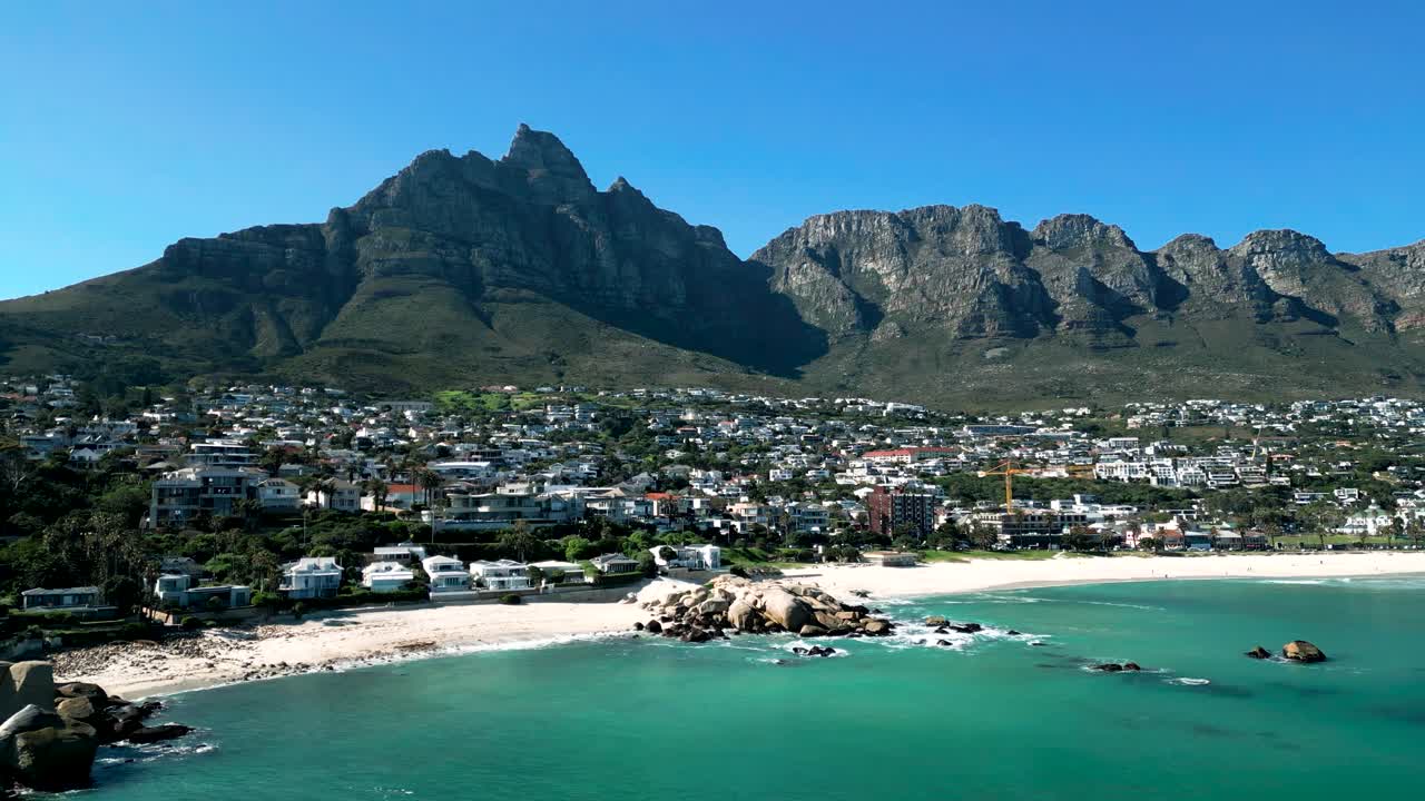 Letecké záběry dronem pláže Camps Bay v Kapském Městě v Jižní Africe s horami v dálce, ukazující pobřežní krajinu a scenérii oceánu.