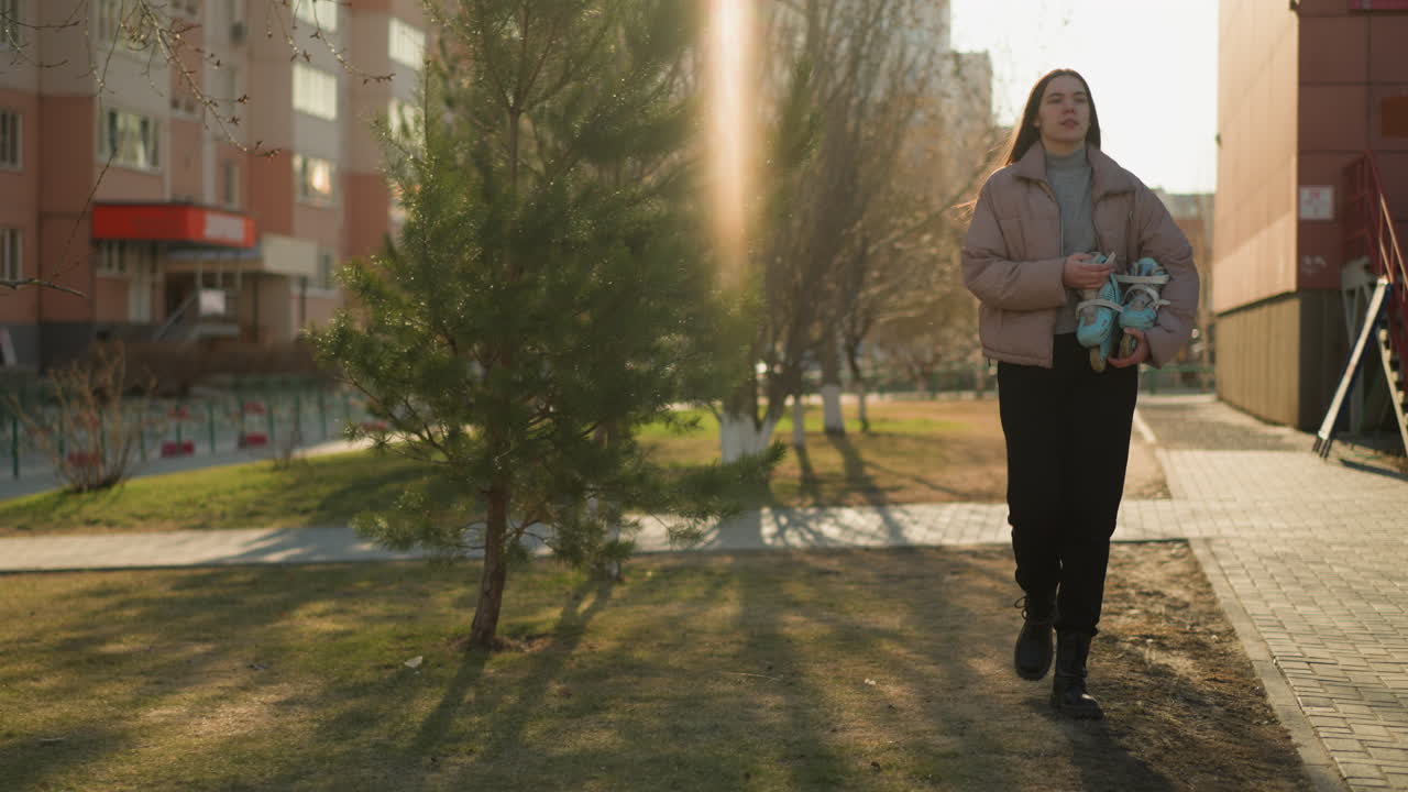 передний вид девушки, держащей скейтборд во время прогулки по городскому району на закате. она одета в персиковую куртку, серую внутреннюю рубашку, черные брюки и черные туфли