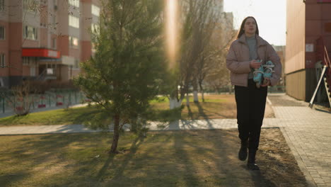 front view of a girl holding a skateboard while walking through an urban area at sunset. she is dressed in a peach jacket, grey inner shirt, black pants, and black shoes