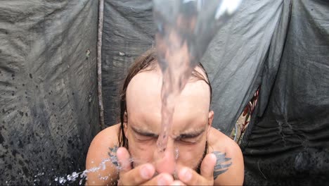 A-young-man-with-long-hair-and-tattoos-is-washing-his-face-and-beard-with-soap-in-a-shower-built-from-sticks,-sheets-of-plastic-and-a-hose