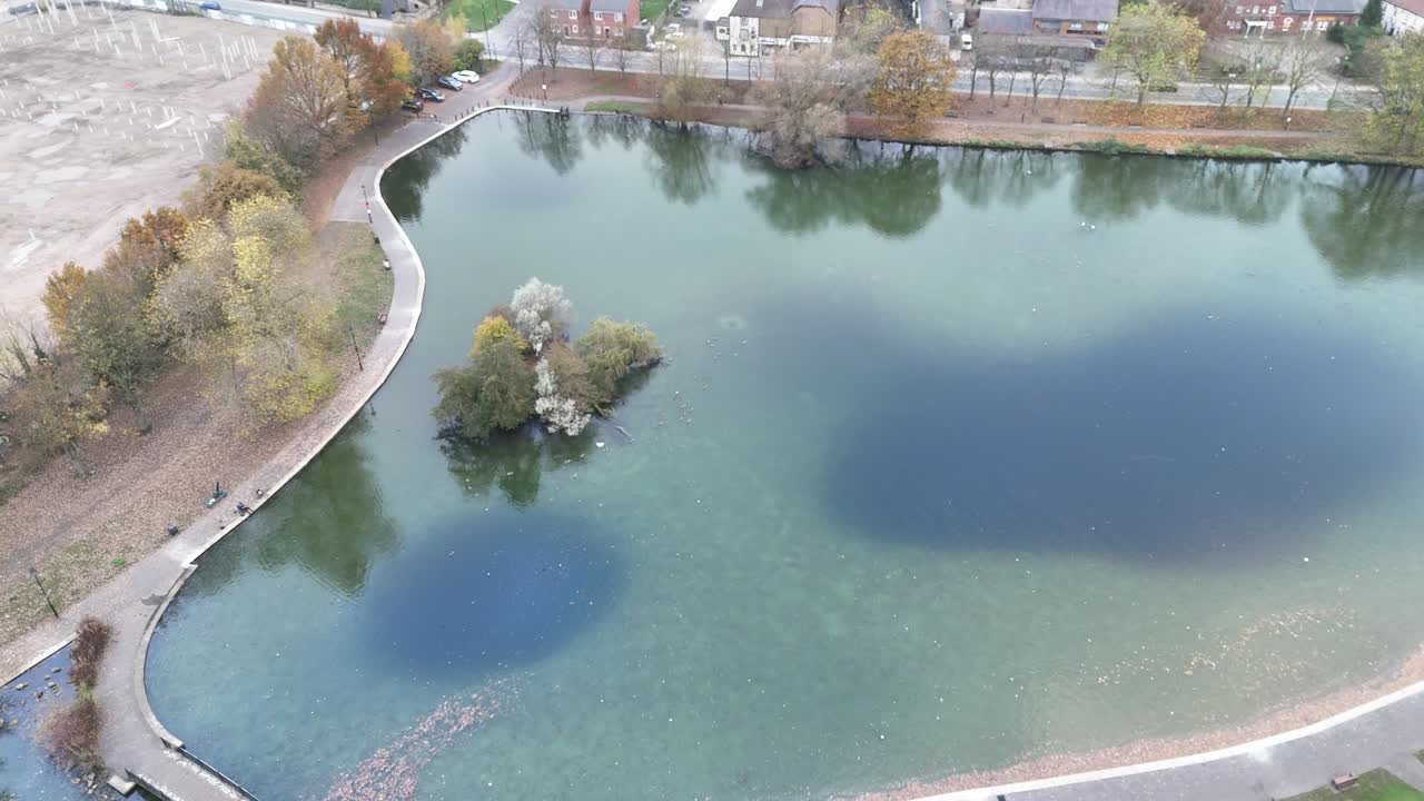 Drone top view of calm pond in Bochum, Germany surrounded by walkway and autumn trees with colorful foliage. Small island with trees sits in clear water showing seasonal beauty and quiet scene