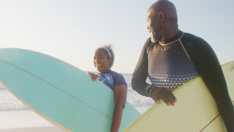 Glückliches-älteres-Afroamerikanisches-Paar,-Das-In-Zeitlupe-Am-Strand-Spazieren-Geht-Und-Surfbretter-Hält