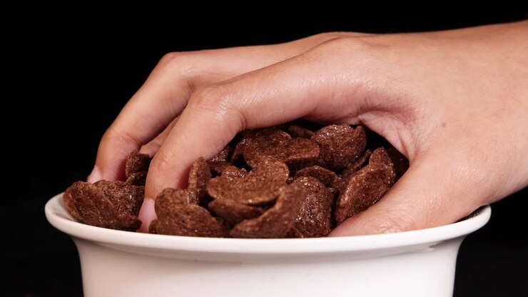 A hand reaches into a bowl of chocolate cereal against a black background, highlighting texture and motion