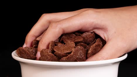 a hand reaches into a bowl of chocolate cereal against a black background, highlighting texture and motion