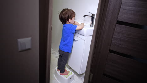 little kid stands on the prop near the sink. cute caucasian toddler washing hands in the bathroom.