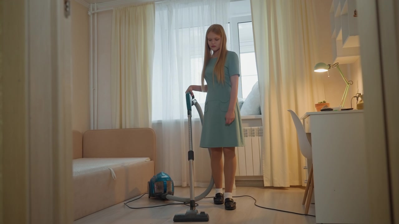 Ung vit kvinna dammsuger vardagsrum, självsäker husägare demonstration, cylinderdammsugare på golvet med sladden slingrad, långsam svepande rörelse över trägolv, ljusa gardiner och soffa, lampdetalj, prydligt.