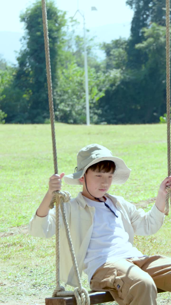 Un bambino che indossa un cappello a secchiello e pantaloni color cachi si diverte a dondolarsi su una corda.