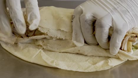 person wearing white gloves rolling grilled food sandwich on stainless table