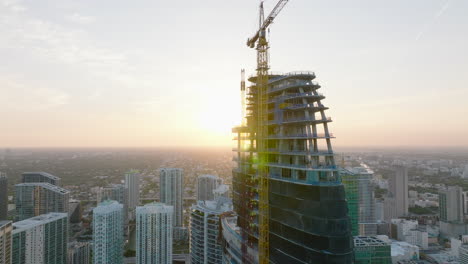 Rising-footage-on-skyscraper-construction-site-against-setting-sun.-Tall-tower-crane-on-top-of-new-building.-Miami,-USA