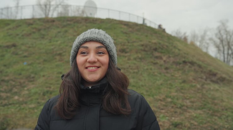 Donna nel parco che balla e batte le mani con un berretto, viso espressivo e sorriso caloroso contro un pendio erboso, il movimento del cappotto invernale mostra improvvisazione e umore allegro sotto una luce nuvolosa