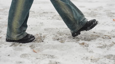 side leg view of someone in black boots and jeans, carrying an item behind them while walking through falling snow, capturing the cold and snowy winter atmosphere