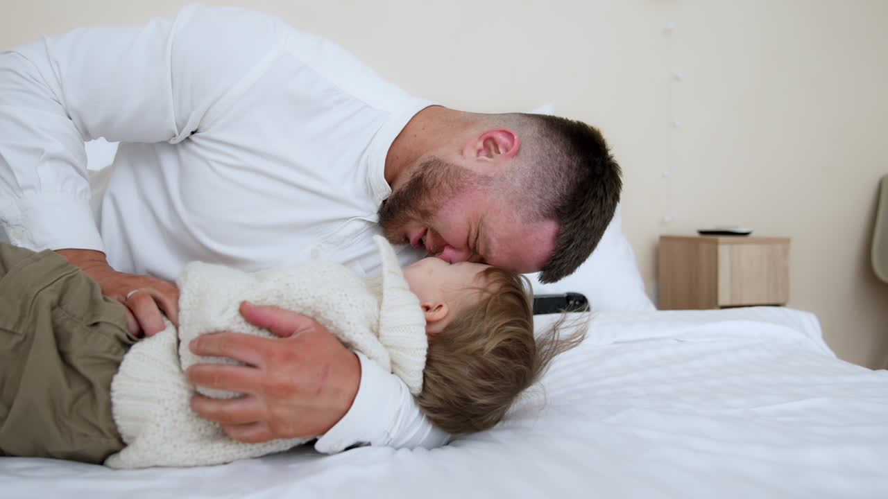 Bearded man hug, kisses and tickles a little cute baby boy on the bed. Loving dad plays with his cheerful son.