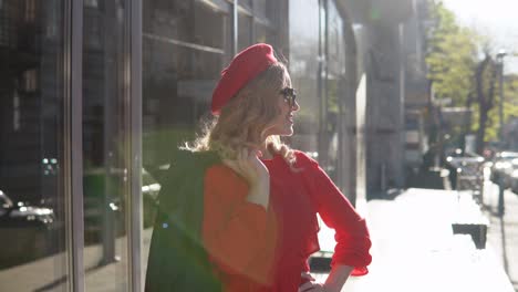 stylish young blonde woman in a red dress and black braid stands on a cozy street in the morning sun