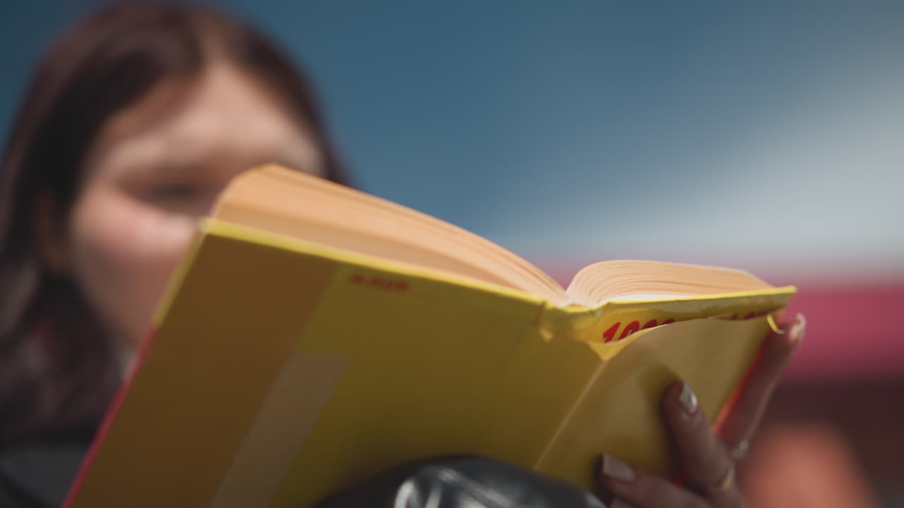 약간 흐릿한 여성의 클로즈업, 그녀의 손에 노란 표지 책의 페이지를 뒤집고, 책의 가장자리와 생생한 색상, 밝은 야외 빛의 배경에 초점을 맞추고