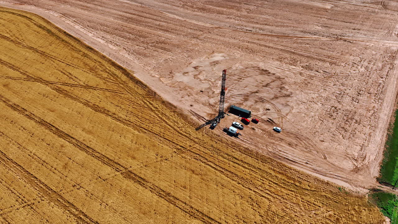 Oil rig with few cars and trucks around. Vast field and wheat plantation around the site. Aerial view.