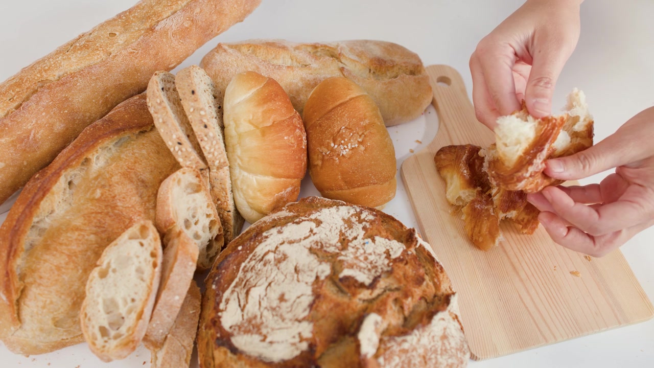 Primo piano di mani che spezzano un croissant sfogliato su un tagliere di legno con vari tipi di pane artigianale