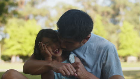 happy asian father sitting with little daughter on the grass and kissing her