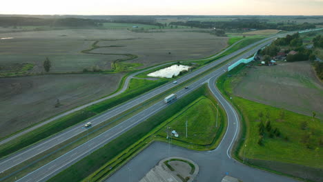 aerial view of a highway with vehicles, surrounded by fields and farmland, with an adjacent parking lot and green areas