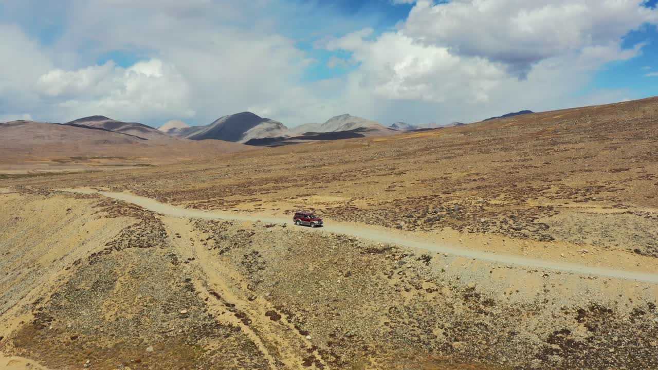 Veduta aerea con drone di un SUV rosso che percorre un sentiero sterrato nell'altopiano alpino arancione del Parco Nazionale Deosai, situato tra Skardu e la Valle di Astore in Pakistan, in una soleggiata giornata estiva