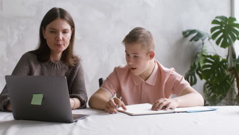 boy with down syndrome watching something on laptop sitting at table with his mother in the living room at home