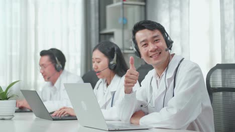 A Man Of Three Asian Doctors With Stethoscope Wearing Headset Working As Call Center Agent Looking Up From Computer Smile Then Thumbs Up While His Colleagues Are Speaking And Typing During A Call At The Office