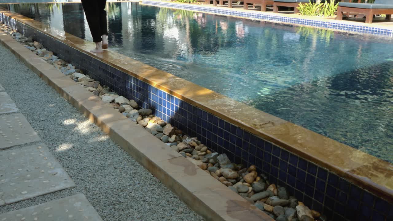 Ung kvinde går langs kanten af en swimmingpool på et tropisk hotel, optaget fra siden