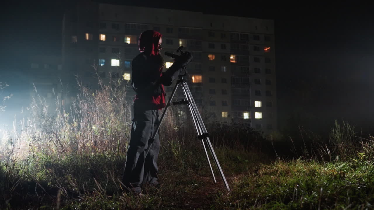 Natlig stjernekigger med teleskop på en mark ved siden af en lejlighedsbygning, hætteklædt observatør justerer stativ og okular midt i tåget græs og lysende vinduer, filmisk svagt lys fremkalder ensomhed og nysgerrighed