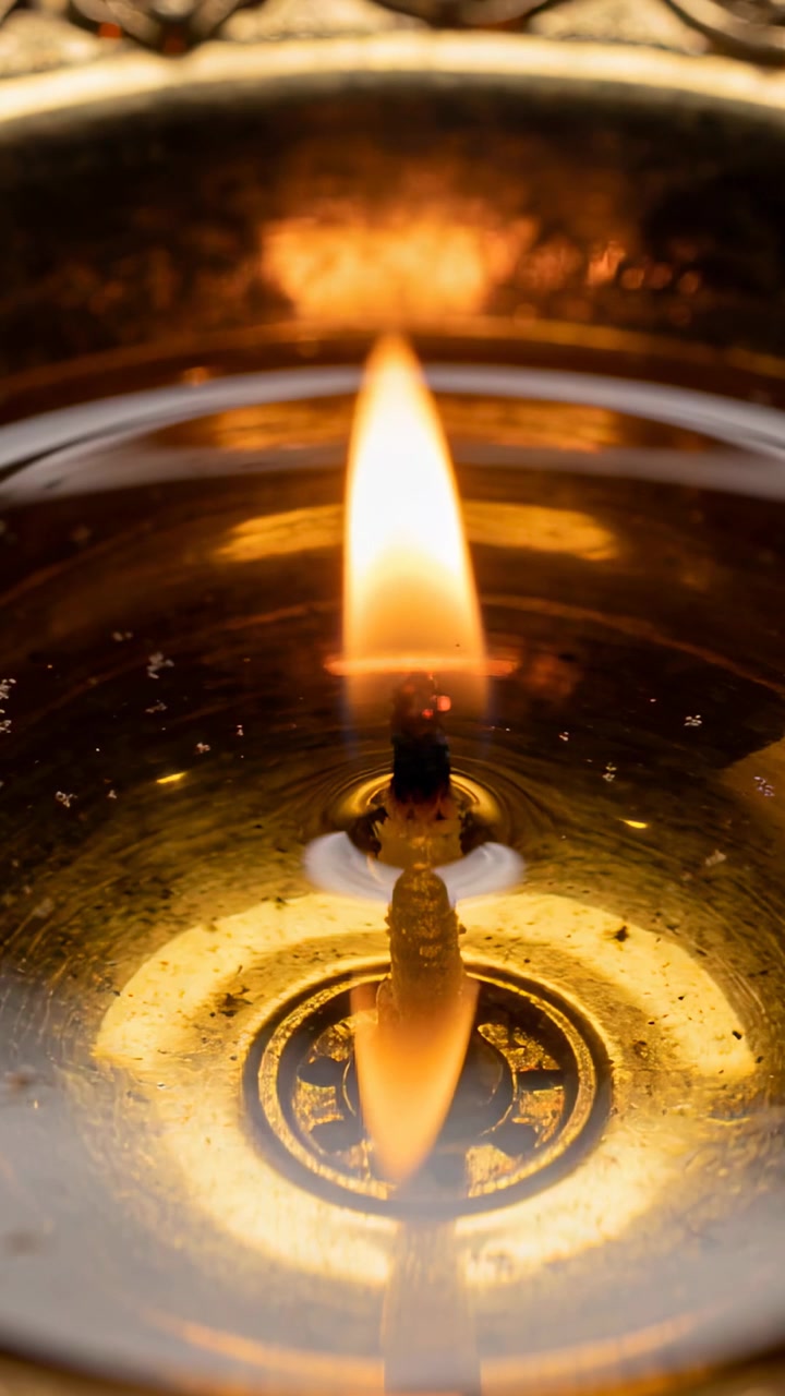 Vertical video: Flickering wick and flame reacting to draft on table in metal bowl mirroring oil