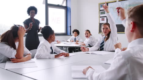 female high school tutor asking pupils wearing uniform sitting at desks question to which they raise their hands
