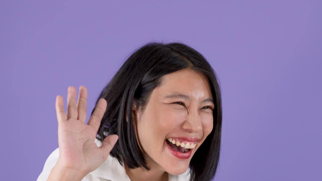 보라색 스튜디오 배경에서 짧은 검은 머리의 쾌활한 아시아 여성이 손을 흔들며 웃고 있어요