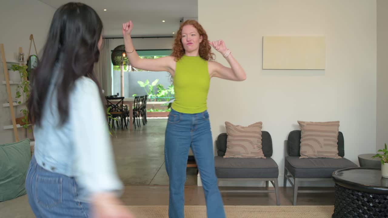 Diverse female friends dancing to beat in home living area by sofa, sharing playful moment