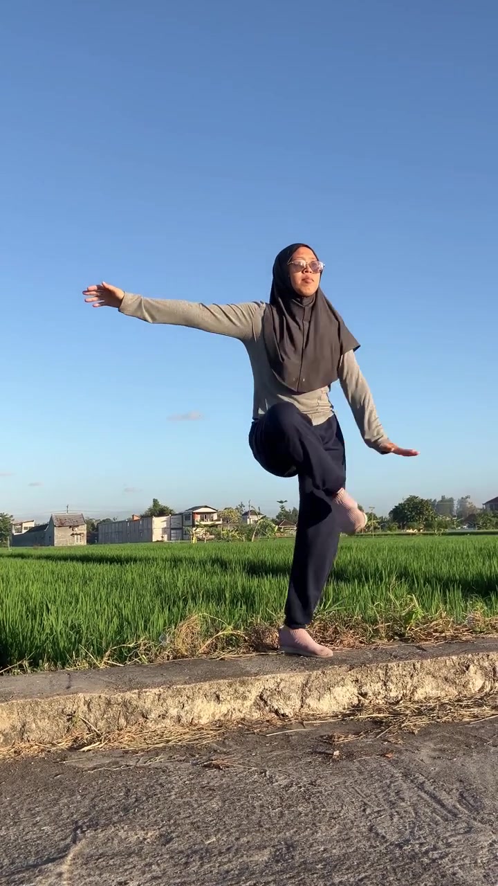 Wanita Muslim muda aktif berhijab melakukan pemanasan keseimbangan satu kaki di luar ruangan di samping sawah hijau yang indah pada pagi yang cerah. Sempurna untuk konsep kebugaran sederhana dan gaya hidup sehat.