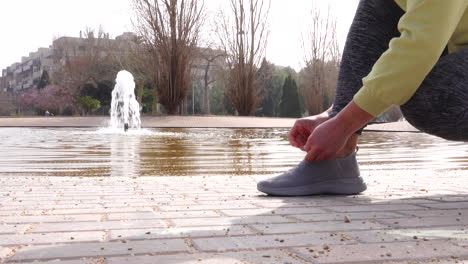 sporty-woman-tying-her-sneakers-in-park-with-a-nice-fountain-in-the-background