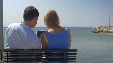 una mujer y un hombre jóvenes están sentados en un banco en la playa en el fondo del horizonte del mar viendo algo en la tableta y hablando en el pireo, grecia.