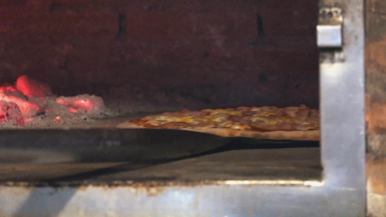 Close Interior Static Shot of a Delicious Large Pizza Being Taken Out of Pizza Oven on Pizza Peel with Red Hot Coals in Pizza Restaurant at Night