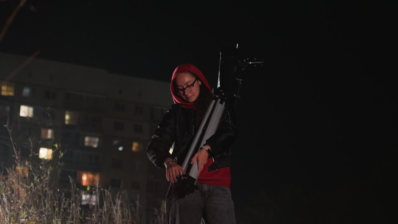 Uno studente regola un telescopio sotto le luci della città di notte, pratica di astrofotografia con treppiede e telescopio accanto a un complesso di appartamenti, figura con felpa rossa che controlla l'allineamento e le impostazioni della fotocamera