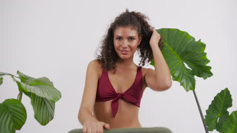 modelo bronceada y de cabello rizado oscuro posando en una silla con un traje de bikini rojo, rodeada de plantas, filmada en un estudio fotográfico