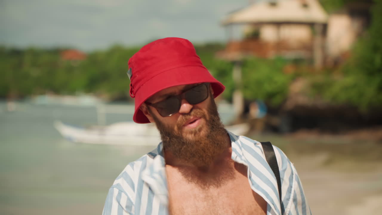 Coppia in spiaggia passeggia con un uomo barbuto, primi piani di sorrisi e conversazioni rilassate mentre l'uomo con cappello da pescatore rosso e camicia a righe trasporta una custodia e uno sgabello vicino a barche e vegetazione, luce calda, fascino marittimo spontaneo.