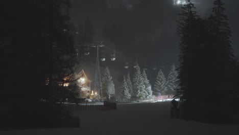 ski gondolas are still in the snowy forest at night, with a warmly lit cabin nestled among tall trees.