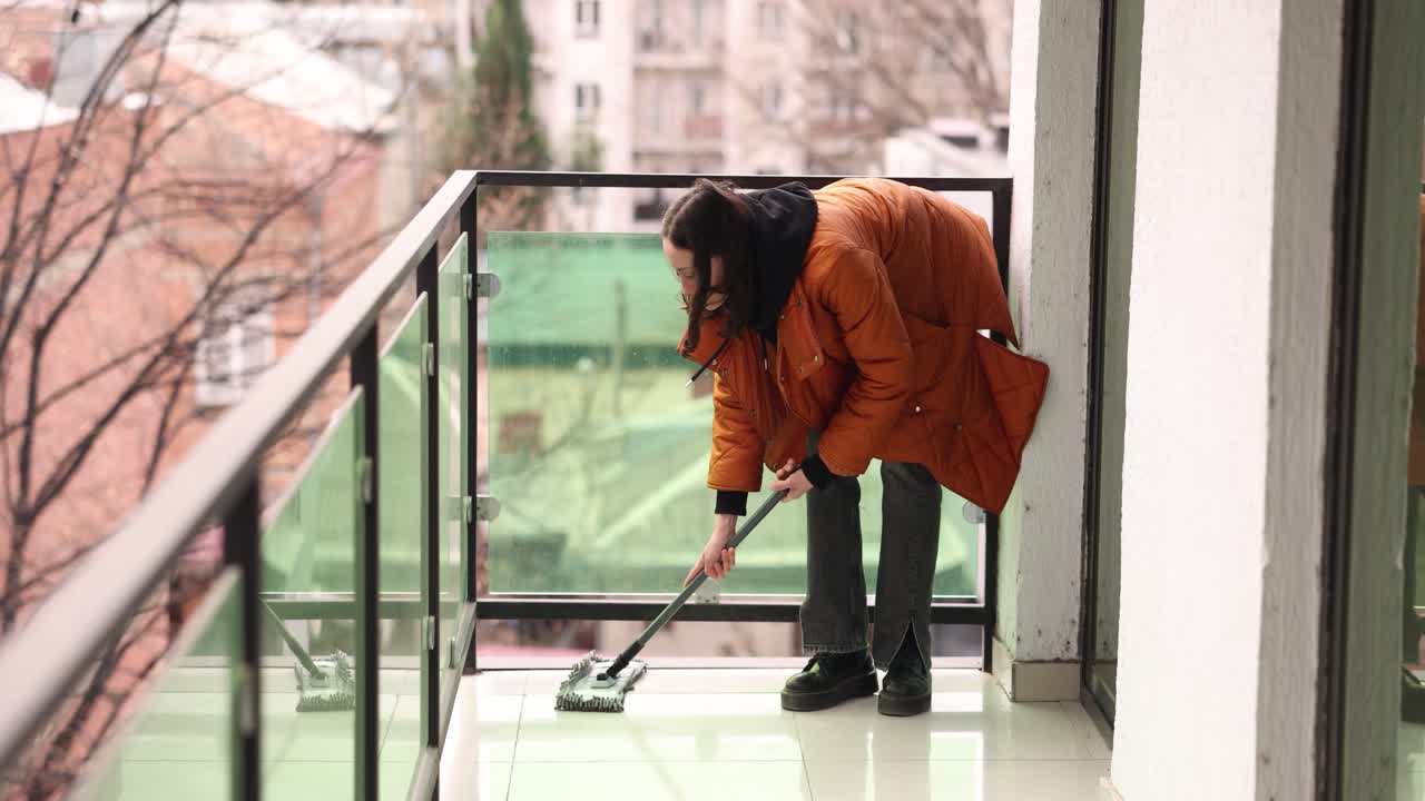 Woman in orange jacket uses mop to clean tiled floor on balcony