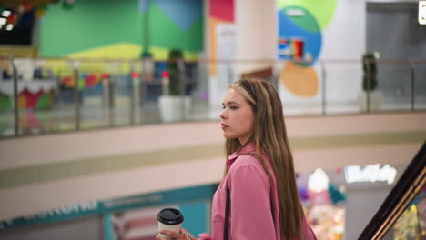 mujer con vestido rosa sosteniendo taza de café y bolsas de compras mientras está de pie en una escalera mecánica mientras desciende en un centro comercial brillantemente iluminado, se pueden ver en el fondo diseños de paredes coloridos y barandillas de vidrio