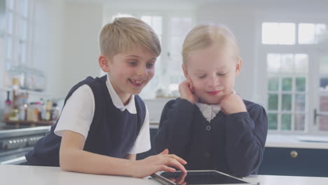 two children wearing school uniform in kitchen playing with digital tablet on counter