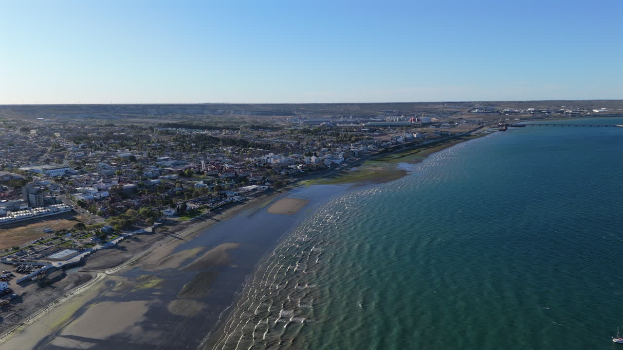 Aerial view of the pier in Puerto Madryn, Chubut Province, Argentina, leading to a vibrant coastal cityscape under a clear blue sky, with the sun reflecting on calm Golfo Nuevo, drone pulling out
