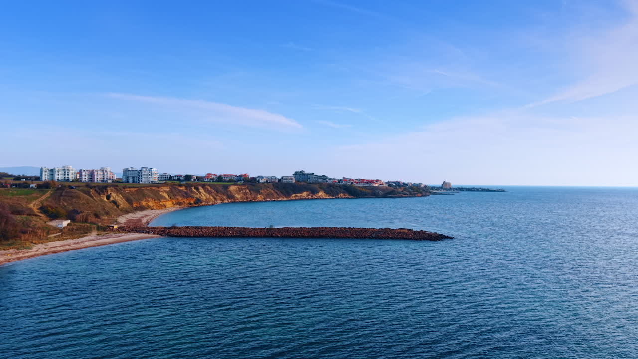 Sereno panorama costiero con scogliere ed edifici sul lungomare. Un tranquillo paesaggio costiero caratterizzato da un mare blu calmo, una spiaggia sabbiosa e scogliere rocciose sormontate da moderni edifici sul lungomare.