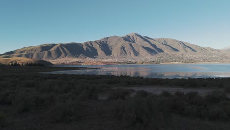 Panorámica-Aérea-Con-Vistas-Al-Agua-En-Un-Paisaje-Desértico-Con-Montañas-Más-Allá-Del-Agua-En-La-Distancia