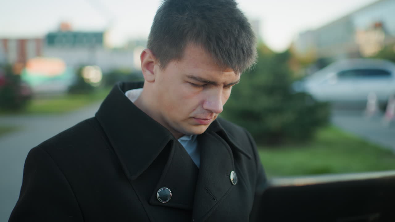 검은색 슈트를 입은 남자가 노트북을 가지고 야외에서 일하고 있으며, 초점을 맞춘 표정, 배경 특징은 주차된 자동차와 도시 건물을 흐릿하게 나타니다.