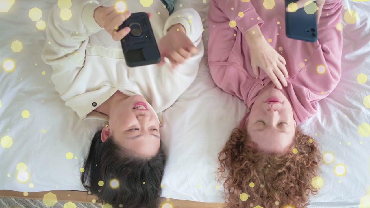 Mulher à esquerda inclinando o telefone, mulheres na cama compartilhando a tela e sorrindo, com uma sobreposição de brilho acentuando a tecnologia.